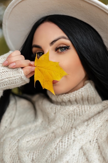 Retrato femenino de otoño de una joven mujer bonita con increíbles ojos azules en un suéter beige vintage y un sombrero de moda que cubre su rostro con una hoja amarilla brillante y mira a la cámara