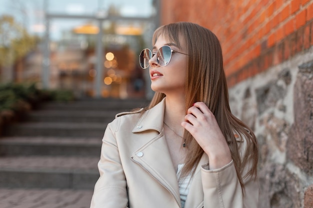 Retrato femenino urbano de una hermosa joven mujer de moda con elegantes gafas de sol redondas vintage en una chaqueta de cuero de moda y camiseta sentada en los escalones cerca de un centro comercial de construcción de ladrillos vintage