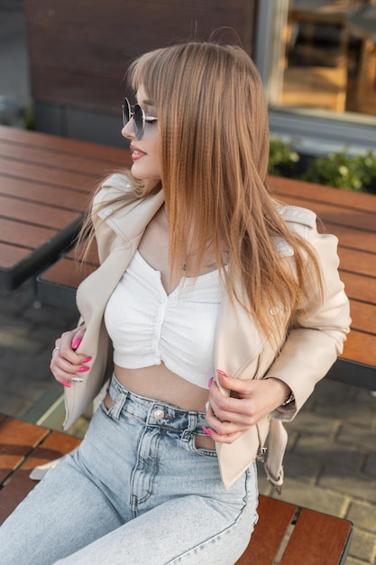 Retrato feminino de moda beleza urbana da bela jovem hipster na moda em roupas de rock streetwear com jaqueta de couro e jeans senta-se no banco de madeira perto de um café na rua