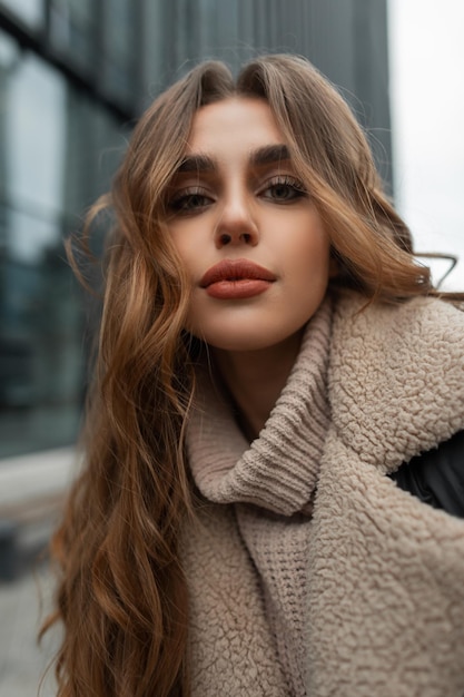 Retrato feminino urbano de um modelo de menina bonita e elegante com um rosto limpo e cabelos cacheados em uma jaqueta de inverno da moda e um suéter de malha vintage na cidade