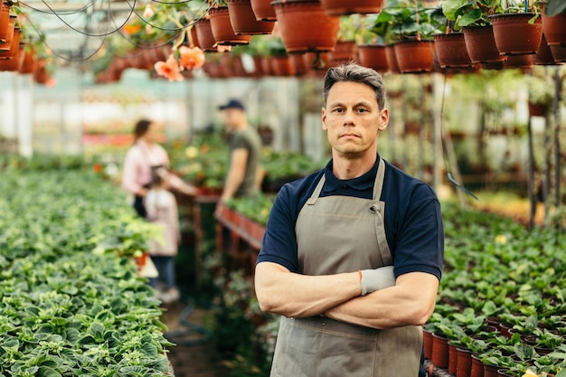 Retrato de floristería masculina con los brazos cruzados de pie en el vivero de plantas y mirando a la cámara