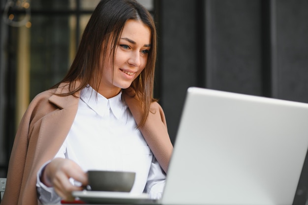 Retrato de la foto de la hermosa mujer atractiva que trabaja en la computadora portátil escribiendo sentado en la cafetería tomando café