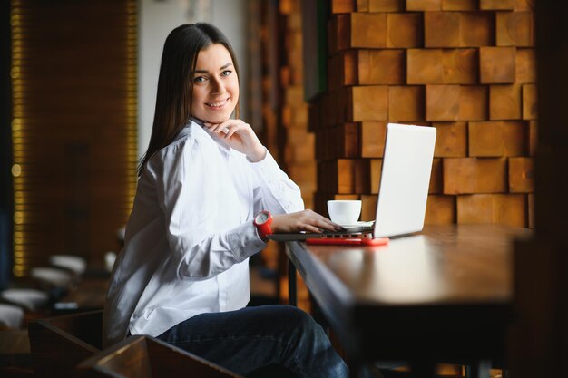 Retrato de la foto de la hermosa mujer atractiva que trabaja en la computadora portátil escribiendo sentado en la cafetería tomando café