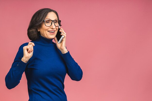 Retrato de la foto de la mujer de edad madura mayor sorprendida sorprendida sorprendida que sostiene el teléfono móvil que ve los descuentos locos vistiendo casual aislado sobre fondo rosado.