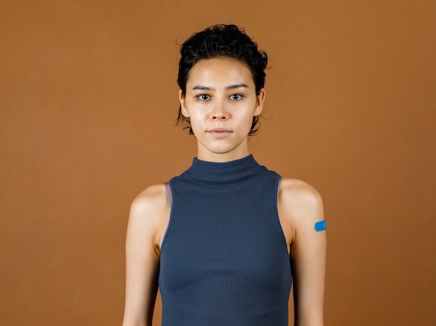 Retrato Foto de la taza del estudio del primer de la paciente femenina atractiva hermosa linda que sonríe mostrando un vendaje de yeso azul en su hombro después de recibir la vacuna contra el coronavirus Covid-19 frente a la pared marrón.