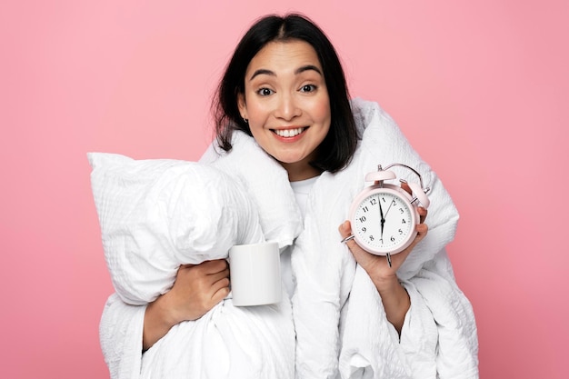 Retrato fotográfico de una chica asiática feliz abrazando una almohada y sosteniendo un despertador