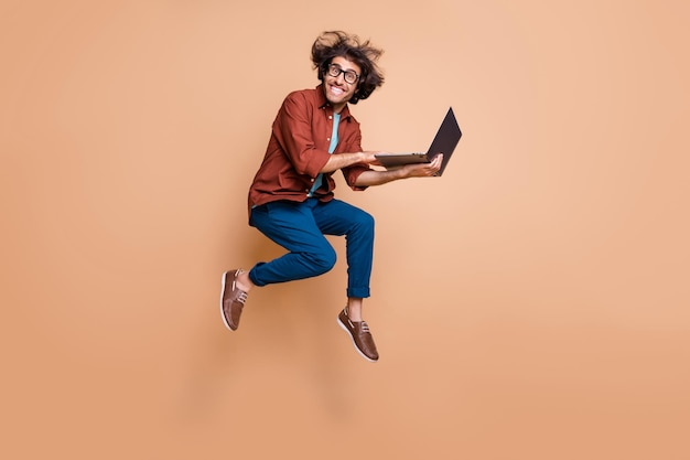 Retrato fotográfico de cuerpo entero de chico emocionado saltando con la computadora portátil en las manos aisladas sobre fondo de color beige pastel