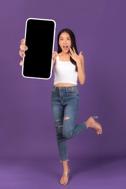 Retrato fotográfico de cuerpo entero de una hermosa joven asiática Emocionada chica sorprendida mostrando un gran teléfono inteligente con pantalla en blanco pantalla blanca aislada sobre fondo púrpura Imagen simulada