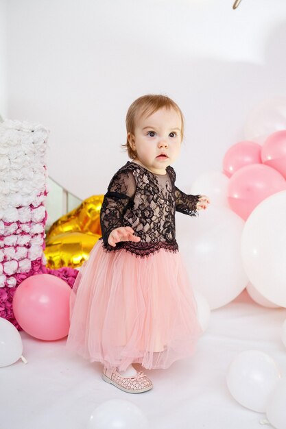 Retrato fotográfico de una cumpleañera de 1 año con un vestido rosa con globos rosas. El niño en la fiesta sonríe, las emociones de los niños. Fiesta de cumpleaños