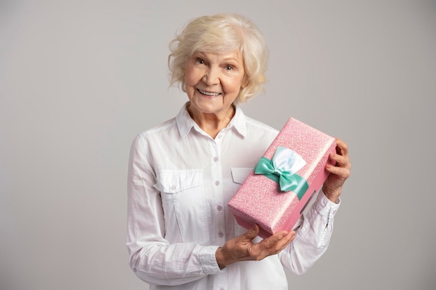 Retrato fotográfico de uma velhinha curiosa segurando caixa de presente e sorrindo isolado em fundo cinza Conceito de dia das mulheres