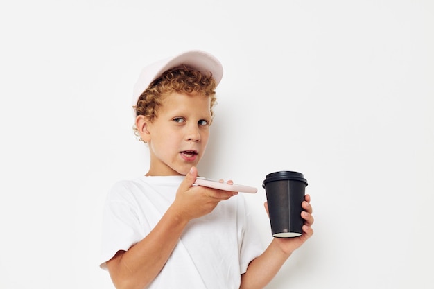 Retrato fotográfico menino encaracolado em um boné de camiseta branca com um telefone em um copo com um fundo leve de bebida inalterado