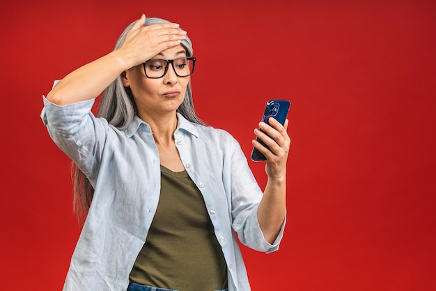 Retrato fotográfico de una mujer madura anciana sorprendida sorprendida asiática sorprendida sosteniendo un teléfono móvil viendo descuentos locos usando ropa informal aislada en un fondo rojo