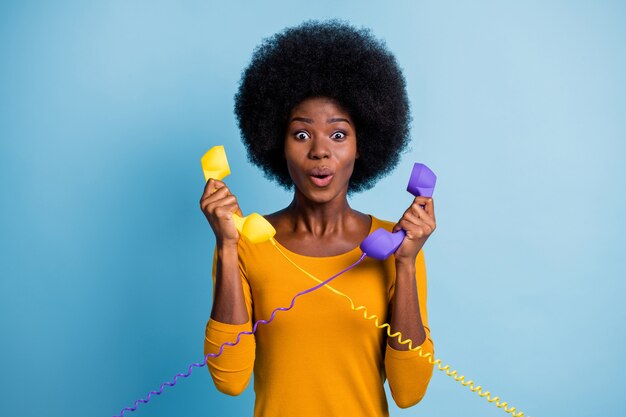 Retrato fotográfico de mujer rizada asombrada de piel negra sosteniendo dos teléfonos retro con cables aislados sobre fondo de color azul vivo