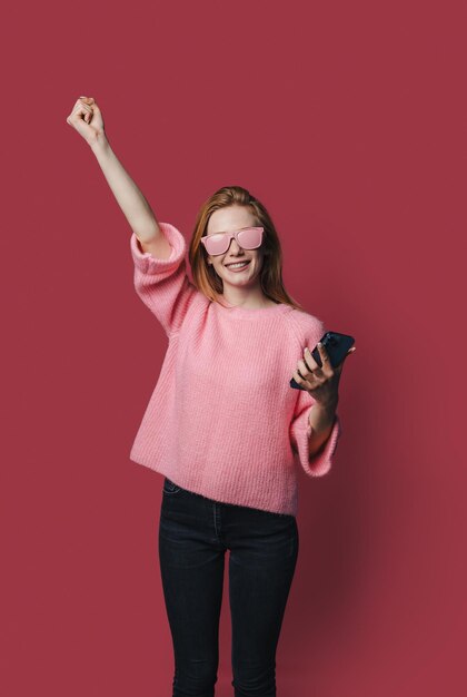 Retrato fotográfico de una mujer sorprendida y emocionada con gafas de sol rosas celebrando sostener el teléfono en una mano aislado en un fondo de color carmesí pastel