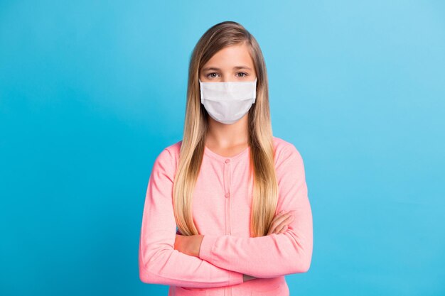 Retrato fotográfico de niña con los brazos cruzados en máscara médica facial aislada sobre fondo de color azul pastel