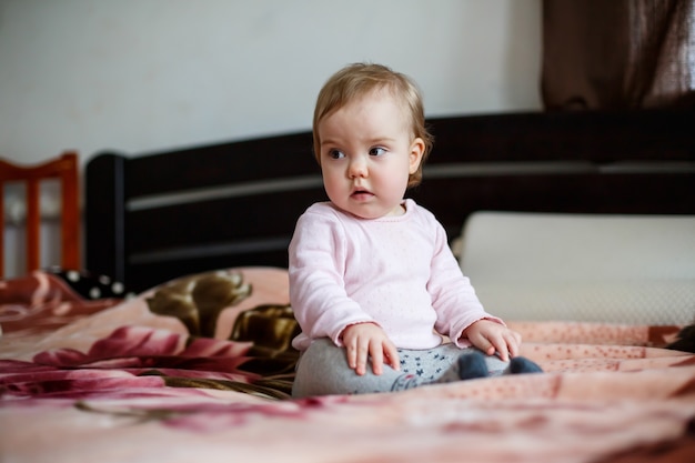 Retrato fotográfico de una niña de mejillas rosadas que se arrastra sobre la cama. Niño sentado en casa