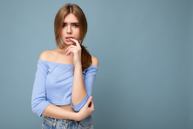 Retrato fotográfico de pensativa joven bonita mujer rubia hermosa con emociones sinceras vistiendo un top azul de moda aislado