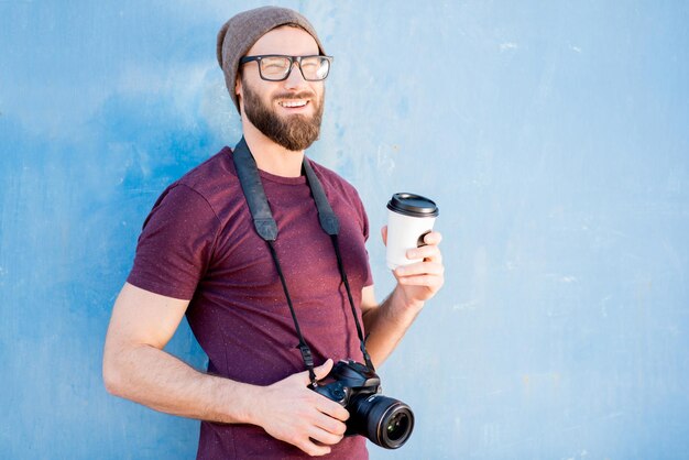 Retrato de un fotógrafo elegante vestido informal con camiseta y sombrero de pie con cámara y café para ir al fondo azul