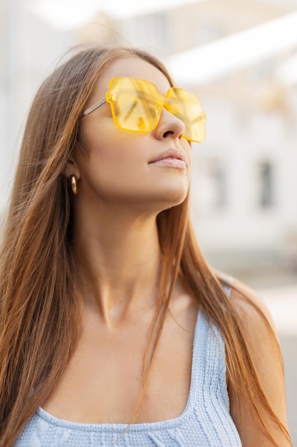 Retrato fresco de una hermosa joven en un traje de verano brillante de moda con gafas de sol amarillas frescas y una camiseta sin mangas de punto azul está caminando en la ciudad