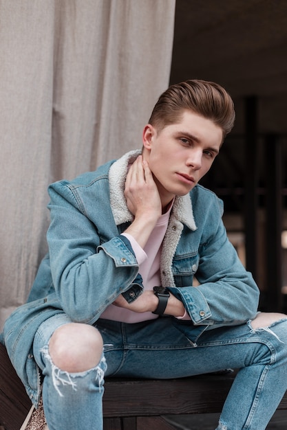 Retrato fresco de moda joven europeo con peinado de moda en elegante chaqueta de mezclilla azul con cuello blanco posando en la calle en la ciudad el día de verano. Chico guapo urbano modelo de moda al aire libre