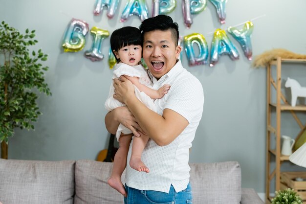 retrato frontal de un alegre padre asiático sosteniendo a su inocente hija y mirando la cámara con una sonrisa con motivo del cumpleaños en casa.