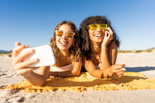 Foto retrato frontal de dos chicas lindas con gafas de sol en vacaciones de verano con smartphone tomando un selfie