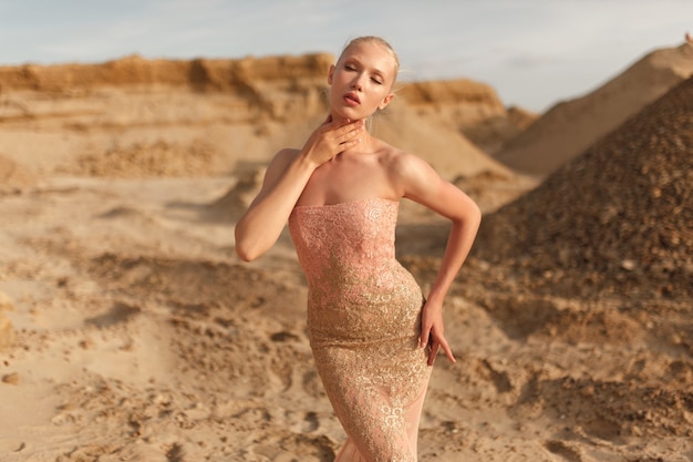 Retrato frontal de una elegante joven vestida con un hermoso vestido ajustado dorado dorado, posando afuera en el desierto al atardecer. Retrato de belleza.