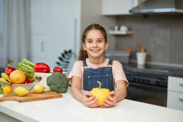 Foto retrato frontal de una linda niña con cabello rizado, vestida con jeans y camiseta, sosteniendo una calabaza amarilla, sentada a la mesa en la cocina.