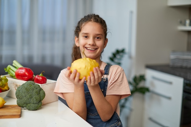 Foto retrato frontal de una niña bonita con cabello rizado, vestida con jeans y camiseta, sosteniendo una calabaza amarilla, sentada a la mesa en la cocina.