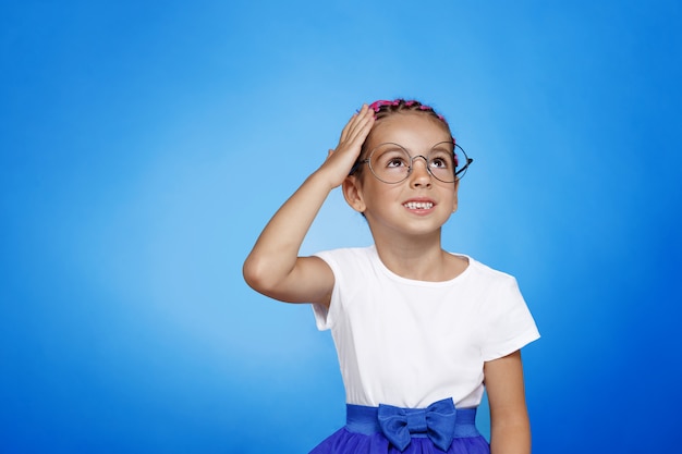 Retrato frontal de una niña bonita confundida en anteojos aislado espacio azul.