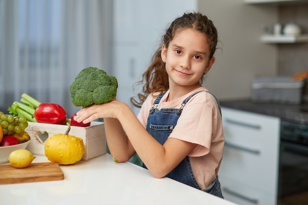 Retrato frontal de una niña con el pelo rizado, sosteniendo y mirando el brócoli, sentado a la mesa en la cocina.