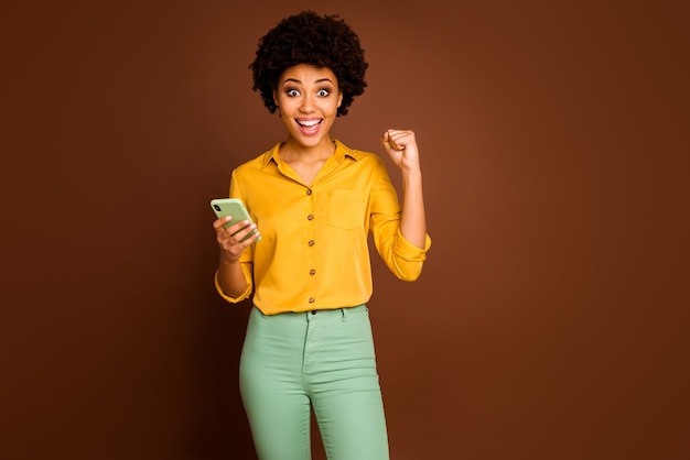 Retrato de funky afortunado afroamericano estudiante blogger uso smartphone celebrar lotería en línea levantar puños gritar sí usar amarillo verde pantalones pantalones color marrón aislado