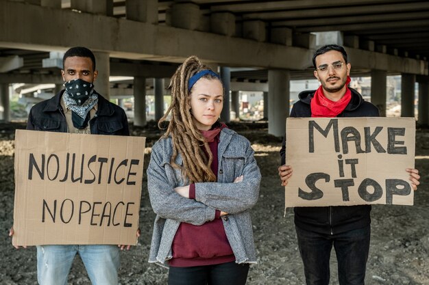 Retrato de gente interracial joven seria de pie con carteles debajo del puente mientras participa en la manifestación