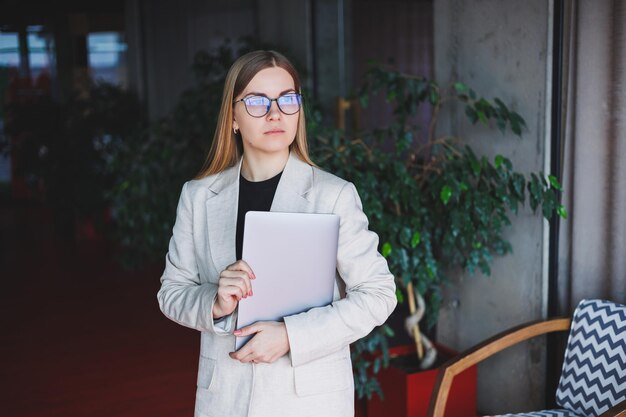 Retrato de un gerente exitoso en la oficina sonriendo durante el día de trabajo mujer feliz con una computadora portátil disfrutando del tiempo en la oficina moderna Mujer rubia con una chaqueta beige y gafas