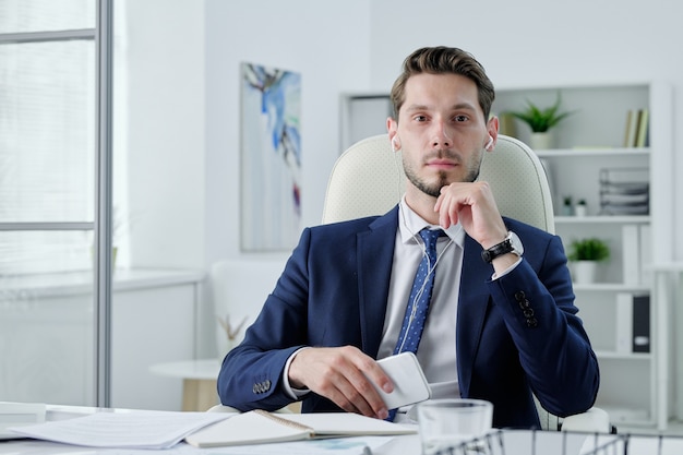 Retrato de gerente de negocios serio en auriculares sentados frente al escritorio y sosteniendo un teléfono moderno en la oficina