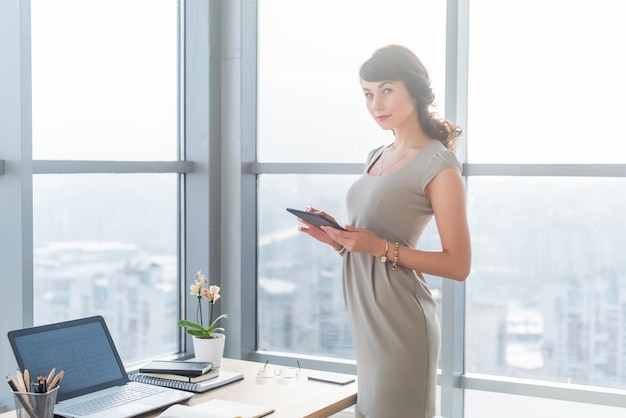 Retrato de una gerente de oficina con un vestido elegante usando su computadora de tableta parada cerca del lugar de trabajo mirando la cámara