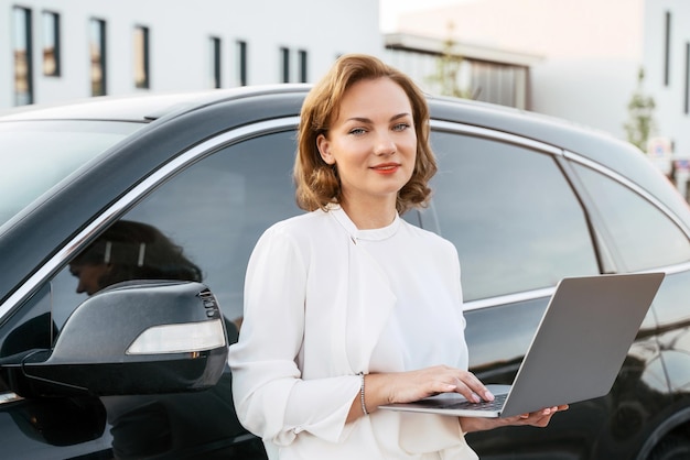 Retrato de un gerente sonriente confiado que usa una computadora portátil cerca de un auto nuevo. Concesionario, alquiler de coches