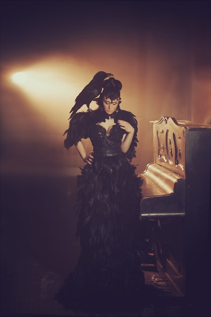 Foto retrato gótico da moda de uma bela morena com corvo em um vestido longo preto feito de penas de corvo. dia das bruxas