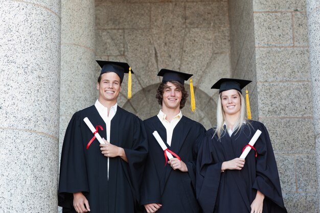 Retrato de graduados sosteniendo su diploma con la universidad en backgroung