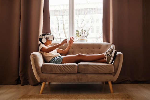 Retrato gráfico de vista lateral de una adolescente negra jugando un juego de vr mientras está acostada en el sofá en casa copiando...