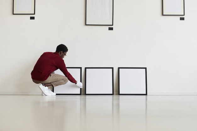 Retrato de gran angular de hombre afroamericano configurando marcos negros en blanco en el piso mientras planifica una galería de arte o una exposición,