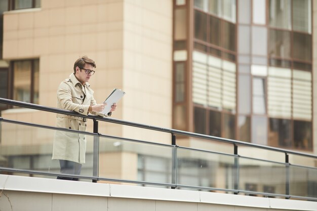 Retrato de gran angular del joven empresario moderno con gabardina y documento de lectura mientras se apoya en la barandilla al aire libre en el entorno urbano de la ciudad, espacio de copia