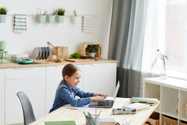 Retrato de gran angular de niña traviesa usando laptop y sonriendo mientras se queda solo para hacer la tarea, espacio de copia