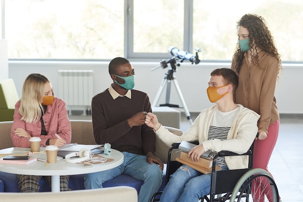 Foto retrato de un grupo multiétnico de estudiantes con máscaras mientras estudiaba en la biblioteca de la universidad con un joven en silla de ruedas en primer plano,