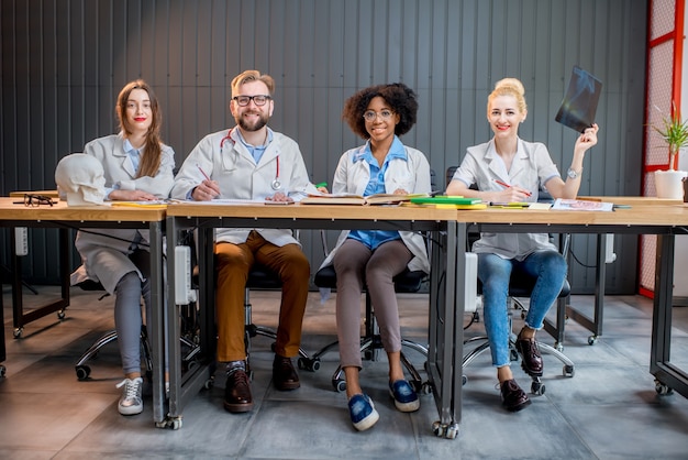 Retrato de un grupo multiétnico de estudiantes de medicina en uniforme sentados juntos en una fila en el escritorio en el aula moderna