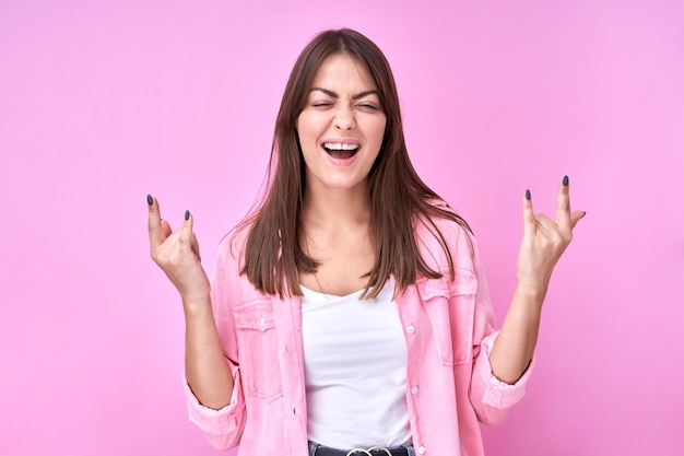 Retrato de una guapa morena hace un gesto de rock n roll que muestra un signo de cuerno de metal pesado que se siente feliz aislado en un fondo rosa