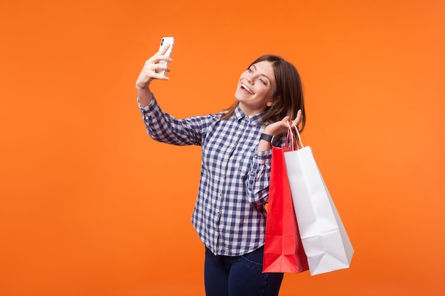 Retrato de una guapa morena con una sonrisa encantadora en un pantalones informal sosteniendo bolsas de compras y hablando de selfie en un smartphone, satisfecha con la compra. tiro de estudio interior aislado sobre fondo naranja