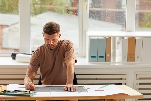 Retrato de guapo arquitecto barbudo mirando planos mientras está sentado en el escritorio de dibujo en la luz del sol,