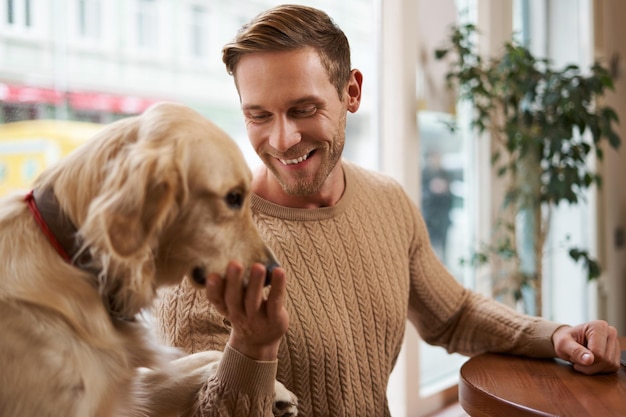 Retrato de un guapo chico rubio sonriente sentado en una cafetería y acaricia a su perro tocando a un golden retriever