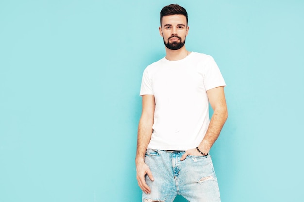 Retrato de guapo confiado con estilo hipster lambersexual modelMan vestido con camiseta blanca y jeans Moda hombre aislado en la pared azul en el estudio